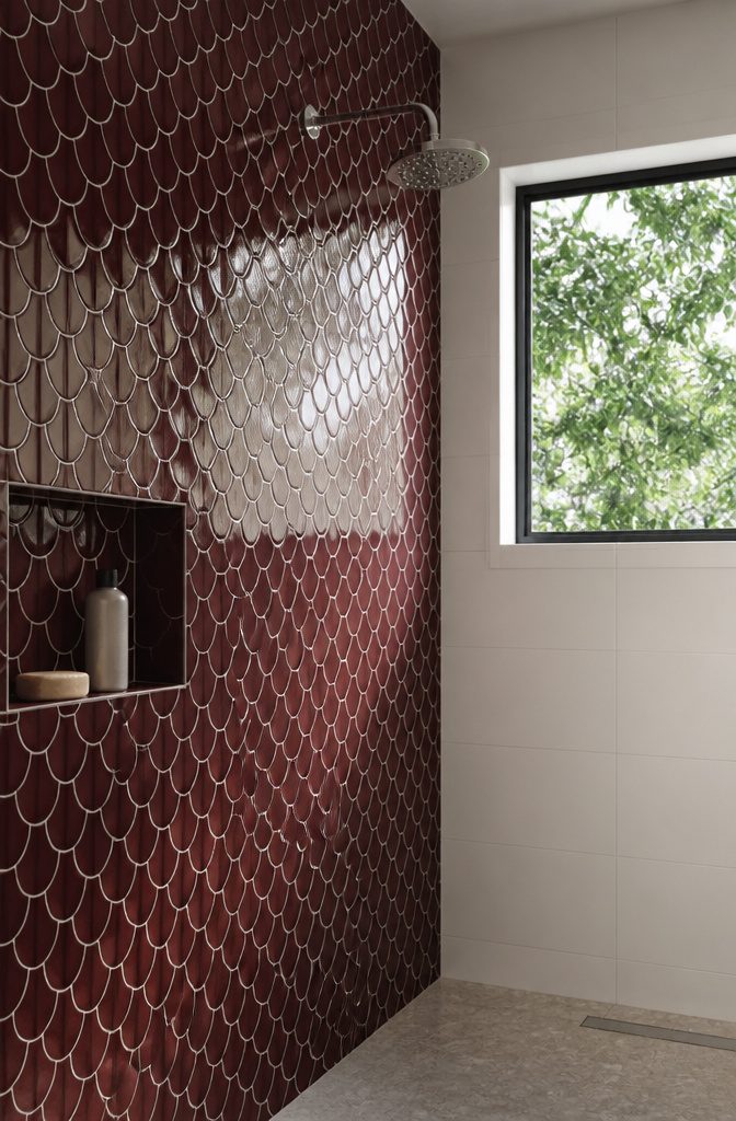 Glossy burgundy fish-scale tiles beside a chrome showerhead and leafy window